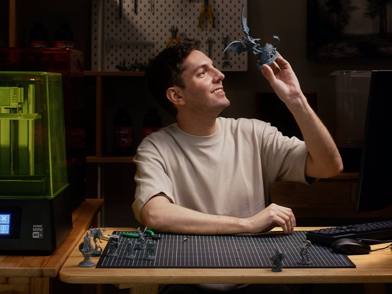 Projetista numa mesa de escritório ao lado da Phrozen Sonic Mini 8K S de tampa verde, olhando uma peça impressa